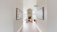 Located at 4901 Meridian Way in Frederick, Maryland, this single-floor community space is thoughtfully designed to blend architectural elegance with modern comfort. The centerpiece of the main area is a dramatic vaulted ceiling that rises to a striking peak, paired with a floor-to-ceiling natural stone fireplace that immediately draws the eye. Expansive multi-pane windows line the walls, flooding the interior with abundant natural light and offering views of the surrounding landscape. Light-toned hardwood flooring runs throughout, complementing the crisp white and warm neutral palette.

The open-plan great room accommodates both a lounge zone — furnished with plush white sofas and accent chairs in warm gold tones — and a defined dining area featuring a solid wood rectangular table surrounded by boucle-upholstered chairs. Pendant lights suspend above the dining space, while recessed lighting illuminates the entire room. A built-in bar counter with open wood shelving and an under-counter beverage cooler provides a convenient refreshment station.

A dedicated office room offers a more intimate setting, with warm taupe walls, crown molding, large windows with garden views, and a round wood table suited for small meetings. The hallway connecting the spaces is accented with eclectic wall art, adding a curated, welcoming character throughout.