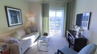 This single-floor space features a well-appointed living area that centers around a large sliding glass door framed by elegant sage-green curtains, which draw in abundant natural light and provide direct access to a private balcony. The balcony offers open outdoor views, adding a sense of spaciousness to the interior. The living area is finished with neutral-toned carpeting that complements the warm, light-colored walls, creating a calm and inviting ambiance. A white sofa with decorative accent pillows anchors the seating arrangement, paired with a sleek, tiered glass and chrome coffee table that reflects the natural light flowing in from the balcony. A dark wood media console and entertainment unit line one wall, providing functional storage and display space. A classic floor lamp with a white shade adds supplementary lighting, enhancing the room's warm, comfortable atmosphere. The overall design balances modern furnishings with a relaxed, airy aesthetic, making the living area feel open and well-connected to the outdoors.