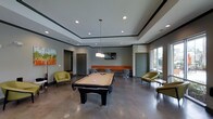 This game room offers a well-appointed recreational space designed for both entertainment and relaxation. At the heart of the room sits a full-size pool table, serving as the central focal point and inviting friendly competition. The polished concrete floors lend an industrial-modern aesthetic that complements the room's contemporary design throughout.

Seating is thoughtfully arranged to encourage socializing, with chartreuse accent chairs positioned throughout the space and an L-shaped bench with orange upholstery nestled along the back wall, creating a casual lounge area. A wall-mounted flat-screen TV above the lounge seating provides additional entertainment options for guests.

The room features a tray ceiling with recessed lighting and a sleek pendant light fixture hanging above the pool table, ensuring ample illumination for gameplay. Large windows along one wall flood the space with natural light and offer views to the outside, enhancing the open, airy ambiance. Colorful artwork on the walls adds a vibrant, welcoming touch to the overall atmosphere. The neutral gray tones of the walls and ceiling are balanced by warm accent colors in the furnishings, creating an inviting and energetic environment well-suited for recreation and community gathering.