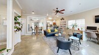 Located at 1515 Aggie Rd in Jonesboro, Arkansas, this single-floor community space is thoughtfully designed to offer both comfort and functionality. The living room serves as the heart of the space, featuring polished concrete floors, crown molding, and ceiling fans that contribute to an open, airy atmosphere. Multiple windows with white blinds flood the room with natural light, offering pleasant outdoor views. The seating area is anchored by a plush area rug and includes a contemporary leather sofa and coordinating accent chairs arranged around a round coffee table — ideal for relaxed gatherings. A built-in workstation along one wall, complete with desktop computers, adds a functional dimension to the room, while built-in shelving and a media console with a wall-mounted television provide additional utility. Lush potted plants throughout lend a warm, welcoming ambiance. A large pass-through opening connects the living area to the kitchen, which features white cabinetry, a granite-topped breakfast bar with upholstered bar stools, pendant lighting, and full appliances including a refrigerator and microwave. Crown molding and neutral warm-toned walls tie both spaces together cohesively, creating an inviting and versatile environment.