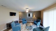 This single-floor space showcases a thoughtfully designed living area that blends comfort and contemporary style. The main living room serves as the centerpiece, featuring rich hardwood-style flooring that contrasts beautifully with the neutral white ceiling and bold charcoal accent wall. A decorative wall mirror with a metallic frame adds depth and visual interest to the room, while a stylish ceiling light fixture provides warm, ambient illumination complemented by a classic floor lamp in the corner. The seating arrangement is anchored by a set of coordinating teal upholstered armchairs with nailhead trim detailing, surrounding a distinctive metallic drum coffee table atop a light-toned area rug. A wall-mounted flat-screen television is positioned opposite the seating area, enhancing the room's functionality as an entertainment and gathering space. A low-profile media console sits beneath the TV, offering practical storage. Natural light filters in through a window, brightening the space alongside the interior lighting. A decorative potted plant adds a touch of greenery, completing the polished, modern ambiance of this inviting living space.