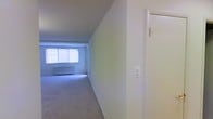 Located in Silver Spring, Maryland, this single-floor space offers a straightforward and functional layout with a clean, neutral aesthetic throughout. The interior is finished with wall-to-wall carpeting that runs seamlessly across the open living area, providing a soft and comfortable underfoot feel. The walls are painted in a crisp, bright white, contributing to the overall sense of openness and light within the space. A large window at the far end of the main living area allows natural light to pour in, illuminating the room and creating an airy atmosphere. The window is fitted with horizontal blinds, offering adjustable light control and privacy. A baseboard heating unit is positioned beneath the window, ensuring efficient climate control throughout the colder months. The layout flows in a long, corridor-like fashion from the entryway toward the main living area, making the space feel elongated and well-defined. A door with standard hardware is visible near the entry, adding to the practical functionality of the unit. The overall ambiance is calm and understated, presenting a blank canvas well-suited for a variety of interior styles and furnishing arrangements.