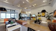 This single-story space is designed as a stylish communal gathering area, combining open lounge, dining, and kitchen zones in a cohesive, contemporary layout. The main lounge area is anchored by an expansive sectional sofa upholstered in a bold mix of orange, yellow, and patterned fabrics, complemented by accent chairs in matching tones. Coffered ceilings with recessed lighting add architectural depth, while large windows dressed in sheer white curtains and Roman shades flood the interior with natural light and offer views of lush outdoor greenery.

A curated gallery of large-format artwork lines the walls throughout, adding visual interest and a creative energy to the space. Light hardwood flooring runs continuously through each zone, unifying the layout. The kitchenette features a long white quartz island with a warm wood accent panel, red leather bar stools, and striking teardrop-shaped blue glass pendant lights above. Dark cabinetry, stainless steel appliances, and a wide pass-through window further define this functional area.

An adjacent dining zone offers round dark-topped café tables with upholstered chairs, set beside floor-to-ceiling black-framed glass doors that open directly to an outdoor area, blending the interior with its tropical surroundings.
