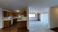 This single-floor residence offers a practical and comfortable layout centered around an open-concept design that connects the kitchen and living area seamlessly. The kitchen is well-appointed with rich wood cabinetry, white appliances including a refrigerator, stove, and built-in microwave, and a peninsula that provides additional counter space and serves as a natural divider between the two areas. The flooring in the kitchen is a smooth hardwood, adding warmth and easy maintenance underfoot. The adjacent living room is finished with plush carpeting, creating a cozy and inviting atmosphere. A ceiling fan is centrally mounted, offering both light and air circulation throughout the space. Natural light filters in through a large window, brightening the room and providing a pleasant view of the outdoors. A sliding glass door with vertical blinds leads to an outdoor area, offering a convenient indoor-outdoor connection. A interior door provides access to additional rooms within the home. The overall ambiance is clean, functional, and welcoming, making it a comfortable everyday living environment.