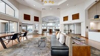 Located at 11260 Point East Dr in Rancho Cordova, CA, this impressive single-floor clubhouse spans a generously sized footprint designed to foster community and connection. The space is defined by soaring double-height ceilings with elegant tray ceiling details and a striking gold multi-globe chandelier that serves as a central design focal point. Warm hardwood flooring in a herringbone pattern flows throughout, complemented by large-scale patterned area rugs that delineate distinct seating and gathering zones. The main living area features plush leather sofas, lounge seating, and a live-edge wood communal table surrounded by bar-height chairs, all bathed in natural light from expansive arched windows with views of the surrounding courtyard and palm trees. A dedicated dining area with a long conference-style table and wall-mounted display screen provides a functional workspace or gathering spot. A built-in bar with dark cabinetry, pendant lighting, and counter seating adds an upscale amenity element. A separate nook with wood-paneled cabinetry and a vending unit offers convenient refreshment access. Throughout, warm wood accent panels, arched architectural details, and high clerestory windows with wood louvers contribute to a refined yet welcoming atmosphere.