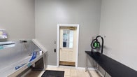 Located at 2201 Williams Glen Blvd in Zionsville, Indiana, this single-floor professional pet grooming facility is thoughtfully designed to provide a clean, efficient, and welcoming environment for both pets and their owners. The interior features neutral gray walls and light tile flooring throughout, creating a bright and hygienic atmosphere that is easy to maintain.

The grooming area is well-equipped with a dedicated stainless steel washing station positioned along one wall, complete with a coiled water hose and grooming supplies within easy reach. Opposite the wash station sits a sturdy black grooming table, accompanied by a wall-mounted professional pet dryer unit in a distinctive green finish, ensuring all essential tools are conveniently accessible in one workspace.

A windowed entry door with a keypad access system provides secure and controlled entry to the space, allowing natural light to filter into the room. The door features classic white trim that complements the clean aesthetic of the interior. A rubber anti-fatigue mat at the entry point adds a practical safety element to the grooming workflow.

The layout is designed with functionality and ease of movement in mind, making it straightforward for groomers to navigate between washing, drying, and grooming stations. The overall ambiance is professional, tidy, and purpose-built for an efficient pet care operation.