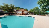Located at 200 Governors Ct in Cartersville, Georgia, this two-floor community facility centers around a generously sized outdoor swimming pool with crystal-clear turquoise water. The pool is flanked by an expansive concrete sundeck that provides ample space for lounging and socializing. The deck is well-appointed with white lounge chairs, seating tables, and vibrant teal market umbrellas that offer shaded areas throughout the day. Mature trees lining the perimeter provide a natural canopy and a sense of privacy, while a wrought-iron security fence encloses the entire pool area. A brick-exterior clubhouse building with a hip roof and chimney anchors one end of the pool deck, providing functional support facilities for pool visitors. Carefully maintained landscaping, including manicured shrubs and decorative plantings, adds color and texture along the fence line. The overall ambiance of the space is bright, open, and welcoming, designed for outdoor recreation and relaxation under the Georgia sun.