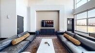 Located at 2020 4th St in Berkeley, CA, this contemporary two-floor communal space offers a thoughtfully designed environment ideal for gathering, working, and socializing. The expansive main floor commands attention with soaring double-height ceilings, exposed industrial ductwork, and rich dark hardwood flooring that anchors the open-plan layout. Floor-to-ceiling gridded windows flood the interior with abundant natural light and frame views of the surrounding landscape, creating an airy, welcoming atmosphere throughout.

The main level is organized into distinct functional zones. A lounge area features sleek black leather sofas centered around a marble-top coffee table and a wall-mounted flat-screen TV set within a recessed architectural niche. Nearby, a dining and collaboration zone offers a large white-top table with industrial-style seating. A decorative laser-cut metal staircase railing serves as a striking artistic focal point, adding visual texture and character to the space.

The kitchen area is outfitted with warm wood cabinetry, white countertops, stainless steel appliances, and pendant globe lighting, providing full functionality for events and gatherings. The upper mezzanine level, accessed by two staircases, features patterned carpet flooring, decorative laser-cut privacy screens, round bistro tables, and built-in wood cabinetry, offering an intimate retreat with panoramic outdoor views.