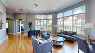 This well-appointed single-story community clubhouse is designed to offer a comfortable and functional gathering environment. The main lounge area is the centerpiece of the space, featuring warm hardwood-style flooring, elevated ceilings accented with crisp crown molding, and recessed lighting throughout. Large floor-to-ceiling windows line the exterior walls, flooding the interior with abundant natural light and offering unobstructed views of an outdoor pool and palm-lined courtyard beyond.

The lounge is furnished with a cohesive collection of blue upholstered armchairs and a gray sofa arranged around a round glass-top coffee table, creating an inviting and relaxed atmosphere. Adjacent to the main seating area, a dedicated dining nook features a round wood table with gray upholstered chairs beneath a stylish drum pendant light, accented by soft blue walls and framed artwork.

A staffed reception area is visible through interior pass-through windows, connecting the lounge to administrative offices. The business center offers a curved granite-topped workstation accommodating multiple computer stations, built-in shelving with arched architectural detailing, and ample natural light from plantation-shuttered windows. The consistent blue-gray color palette and hardwood flooring unify the entire space cohesively.