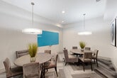 This contemporary dining room offers a well-appointed and versatile gathering space designed with both comfort and functionality in mind. The room accommodates two round dining tables, each surrounded by plush, taupe upholstered chairs — a layout that encourages intimate conversation while efficiently utilizing the available floor space. Underfoot, a striking herringbone-patterned carpet in layered shades of gray adds visual depth and texture to the room. Overhead, a pair of large drum pendant lights with white shades provide warm, even illumination throughout the space, complementing the recessed lighting above. One wall features a bold turquoise dry-erase board, adding a functional and vibrant focal point ideal for collaborative use or presentations. On an adjacent wall, a set of large-scale abstract artworks with swirling blue tones introduces an artistic element that elevates the room's aesthetic. Potted ornamental grasses placed on each table bring a touch of organic warmth to the otherwise modern palette. The walls are finished in a clean, bright white, and a glass-paneled interior door with frosted accents connects the room to adjacent spaces, allowing light to pass through while maintaining privacy. The overall ambiance is polished, modern, and welcoming.