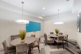 This contemporary dining room offers a well-appointed and versatile gathering space designed with both comfort and functionality in mind. The room accommodates two round dining tables, each surrounded by plush, taupe upholstered chairs — a layout that encourages intimate conversation while efficiently utilizing the available floor space. Underfoot, a striking herringbone-patterned carpet in layered shades of gray adds visual depth and texture to the room. Overhead, a pair of large drum pendant lights with white shades provide warm, even illumination throughout the space, complementing the recessed lighting above. One wall features a bold turquoise dry-erase board, adding a functional and vibrant focal point ideal for collaborative use or presentations. On an adjacent wall, a set of large-scale abstract artworks with swirling blue tones introduces an artistic element that elevates the room's aesthetic. Potted ornamental grasses placed on each table bring a touch of organic warmth to the otherwise modern palette. The walls are finished in a clean, bright white, and a glass-paneled interior door with frosted accents connects the room to adjacent spaces, allowing light to pass through while maintaining privacy. The overall ambiance is polished, modern, and welcoming.