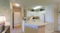 This single-floor residence centers around a bright, functional kitchen designed for both efficiency and everyday comfort. The kitchen is outfitted with a full suite of white appliances, including a refrigerator, electric range, and built-in microwave, all complemented by an abundance of white cabinetry that extends to the upper walls, providing generous storage. Decorative greenery displayed atop the upper cabinets adds a subtle organic touch to the space.

A curved peninsula serves as the kitchen's focal point, offering additional counter space and acting as a natural divider between the kitchen and the adjacent carpeted living area. The peninsula also features a convenient electrical outlet, enhancing its practicality. Recessed and flush-mounted ceiling lighting ensures the kitchen remains well-lit throughout.

Just beyond the kitchen, a paneled entry door with sidelights leads to the foyer, grounding the layout with a clear sense of arrival. Neutral tones in both the cabinetry and flooring create a cohesive, clean aesthetic throughout the visible areas, contributing to an overall atmosphere that is practical, tidy, and comfortable.