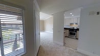 This single-floor space presents a clean, functional layout that connects its main living areas in an open, accessible flow. The transition between rooms is marked by a shift from light-colored carpet in the living and hallway areas to tile flooring in the kitchen, clearly delineating each functional zone. Crown molding runs throughout, adding a subtle architectural refinement to the neutral, warm-toned walls.

The kitchen is well-appointed, featuring white cabinetry that extends to the upper walls, dark granite or stone countertops, and stainless steel appliances including a gas range with an overhead ventilation hood. Recessed lighting in the kitchen ceiling provides bright, even illumination. The tile flooring in this area is practical and easy to maintain.

Adjacent to the kitchen, the carpeted hallway and living spaces offer a softer underfoot experience. A doorway provides access to a secondary room, also finished with carpet and crown molding, maintaining design consistency throughout. Plantation shutters on the windows allow for controlled natural light, contributing to a bright yet private atmosphere. The overall aesthetic is neutral, clean, and move-in ready.