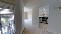 This single-floor space presents a clean, functional layout that connects its main living areas in an open, accessible flow. The transition between rooms is marked by a shift from light-colored carpet in the living and hallway areas to tile flooring in the kitchen, clearly delineating each functional zone. Crown molding runs throughout, adding a subtle architectural refinement to the neutral, warm-toned walls.

The kitchen is well-appointed, featuring white cabinetry that extends to the upper walls, dark granite or stone countertops, and stainless steel appliances including a gas range with an overhead ventilation hood. Recessed lighting in the kitchen ceiling provides bright, even illumination. The tile flooring in this area is practical and easy to maintain.

Adjacent to the kitchen, the carpeted hallway and living spaces offer a softer underfoot experience. A doorway provides access to a secondary room, also finished with carpet and crown molding, maintaining design consistency throughout. Plantation shutters on the windows allow for controlled natural light, contributing to a bright yet private atmosphere. The overall aesthetic is neutral, clean, and move-in ready.