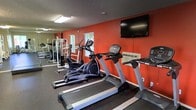 This fitness center is designed to support a well-rounded workout routine in a focused and energizing environment. The space is immediately distinguished by its bold orange accent wall, which lends a motivating, high-energy atmosphere to the room. The dark rubber flooring provides both safety and durability underfoot, making it well-suited for a variety of exercise activities.

The cardio section is prominently featured, housing multiple treadmills and an elliptical machine, each equipped with individual digital consoles for tracking workout metrics. A large flat-screen television is mounted high on the orange accent wall, allowing users to stay entertained during cardio sessions. Natural light filters into the space through windows fitted with horizontal blinds, complementing the overhead fluorescent lighting to keep the room well-lit.

Along the back wall, a white multi-functional strength training rack offers options for resistance and weight training. An additional wall-mounted unit, likely a climate control system, ensures the room remains comfortable during intensive workouts. The textured ceiling adds a classic finishing touch to the space. Overall, this fitness center is a practical, amenity-rich space that caters to both cardio and strength training needs.