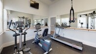 This fitness center is thoughtfully designed to support a well-rounded workout routine within a bright, clean environment. The space is finished with durable rubber flooring throughout, providing traction and cushioning suited for a variety of exercises. Large mirrors line one wall, accompanied by a classic wooden ballet barre, enhancing the sense of space while supporting stretching and balance work. Multiple windows fitted with white plantation shutters allow ample natural light to fill the room, contributing to an open and energizing atmosphere. A wall-mounted television is positioned for easy viewing during cardio sessions. The equipment selection is comprehensive, including a treadmill, recumbent bike, elliptical trainer, and a multi-station cable machine with pulley attachments for strength training. A water cooler is conveniently stationed near the mirrored wall, and a bulletin board displays informational postings for users. The light gray walls and white trim give the space a clean, neutral aesthetic, while the thoughtful layout ensures equipment is accessible without feeling crowded. Overall, this fitness center offers a functional and welcoming environment for daily exercise.