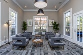 Located at 8064 S Fulton Pkwy in South Fulton, Georgia, this single-floor community space offers a refined and welcoming atmosphere designed for both relaxation and productivity. The interior is finished with warm neutral tones on the walls, crisp white crown molding, and a geometric patterned carpet in cool grays and blues that anchors the entire room with a modern sensibility.

The main lounge area is generously proportioned, featuring a curated arrangement of plush velvet armchairs in a muted blue-violet hue, accented with gold-toned legs and paired with round gold coffee tables. Oversized, white-trimmed windows line multiple walls, flooding the space with natural light and offering views of the surrounding greenery and tree-lined exterior. A bold matte black pendant light fixture serves as a striking focal point overhead, complementing the room's contemporary aesthetic.

A dedicated workstation area is thoughtfully integrated into the space, equipped with a built-in white desk, a wall-mounted flat-screen monitor, and a velvet desk chair, making it a functional work-from-home hub. A glass-paneled French door connects this area to an adjacent space, maintaining visual openness while defining zones. Framed artwork and indoor greenery add warmth and character throughout, completing this polished and practical community environment.