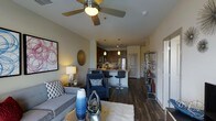 This single-floor residence offers a well-designed open-concept layout that seamlessly connects the living area to the kitchen, creating a cohesive and functional living space. The interior is anchored by rich, dark hardwood flooring that runs throughout, adding warmth and a sense of sophistication to every corner.

The living area is comfortably appointed with a plush gray sofa, a navy accent chair with geometric patterned cushions, and a sleek coffee table adorned with decorative objects. A ceiling fan with integrated lighting adds both practicality and a modern touch overhead, while carefully placed table lamps contribute to a warm and inviting ambiance. Wall art and decorative accents, including a metallic circular piece, lend personality to the space.

The kitchen is a standout feature, boasting dark cabinetry, stainless steel appliances, and a peninsula with bar seating — ideal for casual dining or entertaining. Pendant lighting hangs above the counter, enhancing the kitchen's contemporary aesthetic. A tile backsplash adds texture and visual interest behind the cooking area.

An open metal shelving unit near the entryway provides additional display and storage space. Overall, the residence presents a cohesive, modern design with thoughtful details and a welcoming atmosphere throughout.