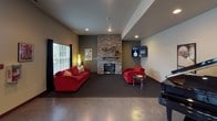 Located at 16900 SE 272nd St in Covington, Washington, this single-story amenity facility offers a well-rounded collection of recreational and social spaces designed for comfort and active use. The building is accessed through a welcoming entryway featuring a bold black double door with decorative glass sidelights, polished concrete flooring, and recessed lighting overhead — setting a clean, functional tone throughout.

The interior corridors connect several distinct areas, decorated with framed artwork and accented by warm beige walls with dark wood trim. A vending area provides convenient refreshment access along the main hallway. The corridor opens toward a spacious lounge area anchored by a striking floor-to-ceiling stacked stone fireplace with a mounted flat-screen television above. Comfortable red seating and a grand piano contribute to the room's inviting, social atmosphere. Natural light filters in through windows dressed with dark drapes.

The fitness center is well-equipped with cardio machines including treadmills and ellipticals, dark carpet flooring, and a full-wall mirror that enhances the sense of space. Rows of windows admit ample natural light, creating an energizing workout environment.

A racquetball court with polished hardwood flooring, clearly marked red boundary lines, and bright white walls rounds out the athletic offerings, providing a dedicated space for high-energy court play.