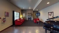 Located at 16900 SE 272nd St in Covington, Washington, this single-story amenity facility offers a well-rounded collection of recreational and social spaces designed for comfort and active use. The building is accessed through a welcoming entryway featuring a bold black double door with decorative glass sidelights, polished concrete flooring, and recessed lighting overhead — setting a clean, functional tone throughout.

The interior corridors connect several distinct areas, decorated with framed artwork and accented by warm beige walls with dark wood trim. A vending area provides convenient refreshment access along the main hallway. The corridor opens toward a spacious lounge area anchored by a striking floor-to-ceiling stacked stone fireplace with a mounted flat-screen television above. Comfortable red seating and a grand piano contribute to the room's inviting, social atmosphere. Natural light filters in through windows dressed with dark drapes.

The fitness center is well-equipped with cardio machines including treadmills and ellipticals, dark carpet flooring, and a full-wall mirror that enhances the sense of space. Rows of windows admit ample natural light, creating an energizing workout environment.

A racquetball court with polished hardwood flooring, clearly marked red boundary lines, and bright white walls rounds out the athletic offerings, providing a dedicated space for high-energy court play.
