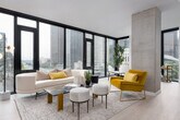 This single-floor residence exemplifies contemporary urban design, characterized by a cohesive, modern aesthetic carried throughout each space. The entry hallway sets a refined tone with light wood flooring, crisp white walls, and curated art, leading seamlessly into the main living areas.

The living room is a standout, dominated by an expansive wall of floor-to-ceiling windows framed in sleek black that flood the space with natural light and offer sweeping city skyline views. The open layout accommodates distinct living and dining areas, maintaining an airy, sophisticated feel throughout.

The kitchen features matte dark cabinetry running the full length of the wall, paired with light countertops, under-cabinet lighting, a matte black faucet, and stainless steel appliances — combining functionality with a sharp, modern look.

The bedrooms each benefit from large black-framed windows with city views. One bedroom features a textured accent wall and carpet flooring, while another incorporates floor-to-ceiling glazing and a bold circular mirror as a design focal point.

Two bathrooms serve the residence: one features marble-tiled tub surround with matte black fixtures, while the other offers a dark vanity with an integrated sink and wall-mounted shelving. Light wood flooring flows consistently across both bathrooms, tying the overall design together.