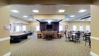 This single-floor community space is designed to accommodate a variety of social activities in a warm and welcoming environment. The expansive main room features a centrally located lounge area anchored by a stone fireplace flanked by rich, dark wood built-in cabinetry and a wall-mounted flat-screen television — creating a natural focal point for relaxation and gathering. Comfortable upholstered seating in warm tan tones surrounds the lounge area, complemented by dark wood accent tables and ottomans.

The room extends generously in all directions, with multiple dining and activity table groupings arranged throughout the space, each furnished with dark wood chairs suited for both casual conversation and structured activities. The patterned wall-to-wall carpeting adds texture and visual warmth to the room while contributing to its acoustic comfort.

Recessed panel ceilings with integrated flush lighting provide bright, even illumination across the entire space. A dropped ceiling detail over the central lounge zone adds architectural definition and a sense of intimacy within the larger room. Warm beige walls and several framed artworks contribute to the inviting atmosphere. Multiple windows along the perimeter allow natural light to fill the space, enhancing its open and airy feel throughout the day.