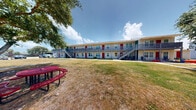 This residential complex is composed of multiple two-story buildings arranged in a courtyard-style layout, creating a communal outdoor environment at its core. The buildings feature a light-colored exterior with distinctive red entry doors and gray railings lining the second-floor walkways, accessible via exterior staircases at each end. The facade design is clean and functional, with each unit appearing to have its own dedicated entry point on both the ground and upper levels.

The central courtyard is a defining feature of the property, offering a generous open-air gathering space anchored by several mature shade trees. Concrete pathways connect the buildings and guide residents through the grounds. A red metal picnic table situated in the courtyard provides a communal seating area ideal for outdoor dining or relaxation. A charcoal grill is also present on the grounds, further enhancing the outdoor living experience for residents.

Surrounding structures include a single-story accessory building with a covered walkway, adding practical utility to the complex. The grounds are primarily grassy, with the mature trees offering significant shade and a sense of established greenery throughout the property. The open sky and abundant natural light give the entire complex a bright, airy character, while the courtyard-centered design fosters a close-knit community atmosphere.