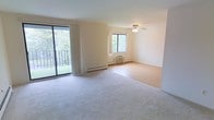 This single-floor living space offers an open, airy layout bathed in natural light from multiple sources. A large sliding glass door dominates one wall, opening onto a private balcony with a metal railing and lush green tree views beyond — a standout feature that brings the outdoors in. Vertical blinds frame the door, providing flexibility in light control.

The room is finished with wall-to-wall plush carpeting in a neutral beige tone, contributing to a warm, comfortable atmosphere. Smooth, light-colored walls throughout keep the space feeling clean and expansive. A ceiling fan is centrally positioned, offering both lighting and air circulation year-round.

A secondary window on the adjacent wall further enhances natural light and ventilation. Climate control is well-equipped, with visible baseboard heating running along the perimeter walls and a wall-mounted air conditioning unit positioned beneath the window — ensuring comfort across all seasons. The overall design is straightforward and functional, offering a versatile, move-in-ready living area with a calm, neutral aesthetic.