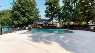 The Tamarind at Apartments community offers a well-appointed outdoor pool area designed for relaxation and recreation. The centerpiece is a generously sized, irregularly shaped swimming pool with clear turquoise water, framed by a broad concrete deck that provides ample space for lounging and movement. The pool is enclosed by a black metal fence with a gated entry, ensuring both safety and privacy for residents.

The surrounding landscape is a standout feature, with large, mature trees providing natural shade and a lush, park-like atmosphere throughout the area. Well-maintained hedgerows and green lawns contribute to the serene setting, while the backdrop of multi-story brick residential buildings adds a sense of community cohesion.

The deck is furnished with outdoor seating, including glass-topped tables and metal chairs, creating comfortable spaces for socializing. A poolside clubhouse or utility structure houses the leasing or management office, identified by a branded entry mat at the door, and features posted pool rules and safety information for residents.

Overall, the pool amenity area exudes a welcoming, community-oriented ambiance, blending practical amenities with a verdant, tree-canopied environment ideal for outdoor enjoyment.