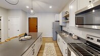 This well-designed single-floor unit offers a thoughtful layout that maximizes functionality across its compact footprint. The open-concept kitchen and living area serves as the heart of the space, featuring white shaker-style cabinetry, dark countertops, open shelving, and a full suite of stainless steel appliances including a refrigerator, electric range, microwave, and dishwasher. A peninsula with seating provides additional workspace and casual dining, illuminated by contemporary globe pendant lights. Light wood-look flooring runs throughout the main living area, complementing the neutral color palette.

The living room benefits from natural light and outdoor access through a sliding glass patio door, creating an airy, open feel. The bedroom is carpeted and features a window with blinds, a baseboard heater, and clean, neutral walls. The full bathroom is finished with wood-look tile flooring, white cabinetry, and Craftsman-style doors with matte black hardware. A dedicated in-unit laundry area houses a full-sized top-load washer and dryer with overhead shelving, adding exceptional practicality to the overall layout.