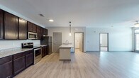 This single-floor unit is designed with a contemporary open-concept layout that seamlessly connects the kitchen and living areas. The kitchen is a standout feature, showcasing rich dark espresso cabinetry with granite countertops that extend to a central island with a built-in sink, illuminated by pendant lighting above. Stainless steel appliances, including a refrigerator, range, and microwave, complete the well-equipped cooking space. Light wood-style flooring runs throughout the main living areas, creating a cohesive and clean aesthetic.

The spacious living area benefits from natural light streaming through double French doors and flanking windows, with a ceiling fan adding both function and style. Neutral gray walls provide a versatile backdrop throughout. Bedrooms feature plush carpeting and large windows that allow natural light and outdoor views.

The bathrooms mirror the kitchen's design language, featuring granite-topped dark vanities with undermount sinks and light wood-style flooring. A dedicated utility room houses the water heater and HVAC system alongside washer/dryer hookups and wire shelving, providing practical storage and laundry capabilities within the unit.