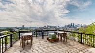Located on Rue Sherbrooke Est in Montréal's vibrant urban core, this rooftop terrace is an elevated outdoor retreat that delivers striking panoramic views of the city skyline, Mont Royal, and the broader metropolitan landscape. The deck features natural wood planking throughout, lending a warm and inviting character to the open-air setting. Black metal railings define the perimeter, providing safety while preserving unobstructed sightlines in multiple directions.

The terrace is thoughtfully arranged with distinct functional zones. A dining area with a rectangular table and chairs offers a comfortable setting for outdoor meals with a backdrop of Montréal's cityscape. Additional lounge seating, including reclining chairs, provides a relaxed space for sun exposure and leisure. Young climbing vines add a touch of greenery along sections of the railing, softening the urban aesthetic.

Rooftop mechanical elements are integrated discreetly into the deck's layout, and a dedicated access door leads directly into the building. The terrace is sheltered on one side by a white brick facade, which provides wind protection while contributing to the architectural character of the space. Under open skies and with sweeping views in nearly every direction, this rooftop terrace presents an exceptional outdoor amenity in the heart of Montréal.