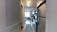 This thoughtfully designed single-floor space blends contemporary style with functional living. The entryway leads directly into a well-appointed galley-style kitchen, characterized by clean white cabinetry, a light wood-toned countertop, and a full suite of stainless steel appliances including a refrigerator, range, and over-the-range microwave. Recessed and flush-mounted linear lighting illuminates the corridor-style kitchen, enhancing the crisp, modern aesthetic. Light hardwood flooring runs throughout, contributing warmth and continuity to the space. A textured runner rug anchors the kitchen walkway, adding a subtle design detail. The kitchen opens seamlessly into a living area that benefits from floor-to-ceiling windows, flooding the space with abundant natural light and offering open city views beyond. The living area is furnished minimally, reinforcing a clean and uncluttered atmosphere. The overall color palette is neutral and contemporary, with white walls, grey tones, and metallic finishes working in harmony. The layout is efficient and well-organized, making excellent use of the available footprint while maintaining a sense of openness and flow throughout the space.