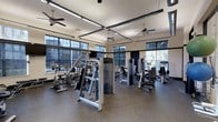 The fitness center is a spacious, single-floor facility designed to support a well-rounded workout routine. The room is lined with large, multi-pane windows on multiple walls, flooding the interior with natural light and offering views of the surrounding outdoor greenery. The generous ceiling height adds to the open, airy feel of the space, while multiple ceiling fans are suspended overhead to maintain comfortable airflow throughout workouts.

The flooring consists of durable, cushioned rubber matting in a speckled finish, providing both safety and support for users. Overhead, the space is illuminated by long, linear fluorescent light fixtures arranged in a grid pattern, ensuring consistent brightness throughout.

The fitness center is outfitted with a diverse selection of equipment, including strength-training cable and resistance machines, stationary bikes, and additional cardio equipment positioned near the windows. The machines are arranged thoughtfully to allow ample circulation between stations. The overall ambiance is clean, functional, and well-maintained, making it a practical and inviting environment for daily exercise.