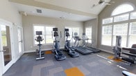 Located at 1431 W Nine Mile Rd in Pensacola, Florida, this single-floor fitness center is designed to offer a bright and functional workout environment. The space is outfitted with a well-curated selection of modern cardio equipment, including stationary bikes, elliptical machines, and treadmills, each equipped with individual display screens for an enhanced exercise experience. Large windows line the walls on multiple sides, flooding the room with abundant natural light and offering views of the surrounding greenery, creating an open and energizing atmosphere. Ceiling fans provide comfortable air circulation throughout the space. The flooring features a durable, patterned carpet tile in neutral tones with contrasting orange accent squares, adding a contemporary visual element to the room. Recessed lighting supplements the natural light for use during all hours. The neutral-toned walls and clean architectural lines contribute to an uncluttered, welcoming ambiance. The layout is thoughtfully arranged to allow ample space between equipment, ensuring ease of movement and a comfortable workout experience for all users.