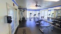 This fitness center offers a bright and functional workout environment designed to accommodate a variety of exercise routines. The space is characterized by large windows that wrap around the room, flooding the interior with natural light and providing views of the surrounding greenery. A ceiling fan mounted at the center of the room promotes air circulation during workouts.

The flooring consists of durable rubber matting, providing cushioning and traction suitable for physical activity. A bold blue accent stripe runs along the upper walls, adding a distinctive visual element that contrasts with the cream-colored walls and crown molding, lending the space a clean and polished aesthetic.

The facility is equipped with multiple cardio machines, including ellipticals, upright exercise bikes, and what appears to be a rowing machine. A multi-station cable weight machine is positioned along one wall, offering options for strength and resistance training. Entry is provided through classic French doors, contributing to the room's refined character. The overall layout allows for comfortable movement between stations, making this a practical and well-appointed fitness facility.