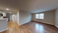 Located at 117 Central St in Acton, Massachusetts, this single-floor residence offers a clean, updated interior with a neutral color palette and consistent design throughout. The layout flows naturally from the entry area through the main living spaces, all finished with warm hardwood-style flooring that adds continuity and brightness to the open areas.

The living room is generously proportioned, featuring large windows that invite natural light and baseboard heating. It connects seamlessly to a dedicated dining or secondary living area, providing flexibility in how the space is used. Two entry doors are visible from this zone, offering convenient access points.

The kitchen is well-appointed with crisp white shaker-style cabinetry, light countertops, and a full suite of stainless steel appliances including an electric range, over-the-range microwave, and dishwasher. A breakfast bar-style partition separates it from the adjacent living area, enhancing the open feel.

The bedrooms feature soft carpeting, multiple windows with blinds for natural light and privacy, and baseboard heating. Their neutral tones create a calm, comfortable atmosphere.

The bathroom is bright and functional, complete with a white vanity, tub/shower combo, and a window for natural ventilation. An adjacent linen closet with built-in shelving adds practical storage to the space.
