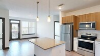 This well-designed 2-bedroom, 2-bathroom residence in Augusta, GA offers a thoughtful layout that maximizes comfort and functionality. The open-concept kitchen is a standout feature, showcasing warm wood cabinetry, white lower cabinets, a classic subway tile backsplash, and a full suite of stainless steel appliances including a range, microwave, dishwasher, and refrigerator. A central kitchen island with pendant lighting connects seamlessly to the living area, which is bathed in natural light from large windows with views of the surrounding area. Light hardwood-style flooring extends throughout the main living spaces, adding warmth and continuity to the design. Both bedrooms feature plush carpeting and ceiling fans, with the secondary bedroom offering dual windows that frame outdoor views. Each bedroom is served by its own dedicated bathroom — one featuring a glass-enclosed shower with a modern vanity, and the other with a full soaking tub and tiled surround. The walk-in closet provides generous wire-shelf storage. A connecting hallway ties all rooms together efficiently, completing this clean and contemporary living space.