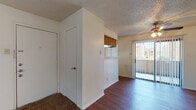 This single-floor unit offers a practical and functional layout with a variety of flooring finishes and an open connection between the main living spaces. The living area is bright and airy, featuring rich hardwood-style flooring, a ceiling fan with light fixture overhead, and a textured ceiling. A full-length sliding glass door with vertical blinds leads to an outdoor balcony, allowing natural light to fill the room and providing outdoor views. A half-wall pass-through connects the living area to the kitchen, which features warm wood cabinetry, a white electric range with overhead ventilation hood, a full-size refrigerator, and light-toned countertops. The galley-style kitchen layout is efficient and well-equipped, with hardwood-style flooring consistent with the living area. Adjacent to the kitchen and living space is a dedicated storage or utility area with a closet door. The bedrooms are carpeted, offering a quieter and softer underfoot feel, with smooth walls and ample space. A separate carpeted room features a window that brings in natural light and an outlook to the surrounding exterior. Throughout the unit, a consistent neutral color palette on the walls creates a clean, move-in-ready atmosphere.