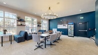 Located at 3227 S Parker Rd in Denver, CO, this single-floor community lounge space offers a welcoming and thoughtfully designed gathering area. The open-plan living room is bathed in natural light, courtesy of expansive white-framed windows that line the walls and offer outdoor views of the surrounding grounds. Soaring ceilings with recessed lighting enhance the airy, open atmosphere throughout.

The space is furnished with a mix of modern seating arrangements, including navy blue lounge chairs and sofas paired with sleek tables, as well as accent chairs in warm wood tones — creating distinct zones for relaxing or casual conversation. A light, textured carpet anchors the interior, complementing the neutral warm-white walls.

A standout feature is a bold teal accent wall adorned with five decorative snowboards showcasing Colorado mountain imagery, nodding to the region's outdoor culture. An adjacent fireplace adds a cozy touch. Floating wooden shelves display curated décor items including plants, books, and art pieces, adding warmth and personality.

A secondary area features deep teal walls with nature-themed artwork and a branded refreshment vending unit, rounding out the space's amenities. Overall, the lounge strikes a balance between modern design and Colorado-inspired character, making it an inviting communal retreat.