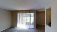 This single-story space presents a straightforward and functional layout. The main living area is defined by neutral-toned walls with a textured ceiling, lending a subtle visual depth overhead. Carpeted flooring runs throughout the room, providing a soft, warm underfoot feel that complements the earthy wall tones.

A prominent feature of the living area is the large sliding glass door fitted with vertical blinds, which serves as the primary source of natural light. The door opens directly to an outdoor area, where lush greenery and open lawn space are visible, creating a pleasant connection between the interior and exterior. The generous glazing allows sunlight to pour into the room, brightening the otherwise neutral interior palette.

The space maintains a clean, open feel with minimal architectural ornamentation. Baseboards line the perimeter of the room, and a single electrical outlet is visible along the lower wall, reflecting a practical, utilitarian design approach. Overall, the space offers a calm, no-frills environment with its most notable asset being the natural light and outdoor access provided by the sliding glass door.