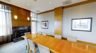 This sophisticated conference room is designed to facilitate professional meetings in a refined, comfortable setting. At its center sits a generous hardwood conference table with a rich, warm finish, surrounded by elegantly upholstered chairs in a neutral woven fabric — providing both style and comfort for extended gatherings. The room's architectural design is defined by warm wood panel accents that line the walls, adding a sense of depth and executive character to the space. A striking dark marble fireplace anchors one corner of the room, serving as a distinguished focal point that elevates the overall ambiance. Recessed lighting in the white ceiling provides clean, even illumination throughout, complemented by natural light filtering in through windows that offer views to the exterior. Wall-mounted artwork adds a touch of curated sophistication. The overall palette — combining soft grey walls, warm wood tones, and dark stone — strikes a balance between welcoming warmth and professional formality, making this space well-suited for boardroom meetings, private discussions, or formal presentations.