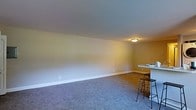 This single-floor space presents a clean, open layout designed for comfortable everyday living. The main living area is generously proportioned, featuring wall-to-wall carpeting in a neutral gray tone that adds warmth and softness underfoot. Smooth, light-colored walls complement the overall understated aesthetic, while white baseboards provide a crisp finishing detail throughout.

Overhead, a flush-mount ceiling light provides functional illumination, keeping the space bright and practical. The room flows naturally toward a kitchen bar area, which is visible along one edge of the space. The bar counter, paired with sleek bar stools, creates a casual dining or socializing spot that connects the living and kitchen zones seamlessly.

The open-concept design allows for flexible furniture arrangements, making it easy to personalize the space to suit a variety of needs. The minimal, neutral palette throughout creates a calm and adaptable ambiance, serving as a blank canvas for any interior style. Overall, this single-level space combines practical design with an approachable, welcoming atmosphere.