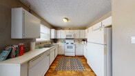 This two-floor residential space offers a functional and comfortable layout with consistent design elements throughout. The kitchen is designed in an efficient U-shape configuration, featuring white cabinetry that extends to the ceiling for ample storage, white appliances including an electric range, built-in microwave, dishwasher, and refrigerator, complemented by light countertops and a double basin sink positioned beneath a window. Warm wood-look flooring runs throughout the space, adding a cohesive and inviting feel. The living room makes a strong visual statement with a bold olive-green accent wall, ceiling fan, and light wood-tone flooring. It offers a relaxed seating arrangement anchored by a wooden coffee table. Two bedrooms provide comfortable retreats, each with textured ceilings, ceiling fans, and wood-look flooring. One bedroom features warm taupe walls, while the other carries an earthy olive tone. A dressing area adjacent to one bedroom includes natural light from a window, a wall mirror, and built-in closet storage. The full bathroom is appointed with a combined tub and shower, white vanity cabinetry, and overhead storage, maintaining the clean, neutral aesthetic found throughout the rest of the space.