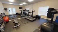 This single-floor fitness center is designed to support a comprehensive workout experience. The space is outfitted with a selection of cardio equipment, including a treadmill, elliptical trainer, and a rowing machine, catering to a variety of aerobic exercise preferences. A dedicated free weight area features a weight bench and a set of dumbbells, providing options for strength training. Rubber flooring and exercise mats are positioned throughout the room, offering cushioning and safety during workouts. A large wall-to-wall mirror spans one side of the room, enhancing the sense of space while allowing users to monitor their form. A wall-mounted flat-screen television is positioned above the mirror, adding an entertainment option during exercise sessions. Natural light filters in through multiple windows fitted with blinds, creating a bright and welcoming atmosphere. The neutral-toned walls and textured ceiling contribute to a clean, practical aesthetic. Overall, the fitness center is thoughtfully arranged to maximize usability and comfort for a range of fitness activities.