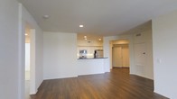 This single-floor space features an open-concept layout that integrates the living area and kitchen seamlessly. The living area is characterized by rich, dark hardwood flooring that extends throughout, providing a warm and grounded aesthetic. Recessed lighting is installed throughout the ceiling, offering even, ambient illumination across the space.

The kitchen is defined by a wraparound peninsula counter with a dark countertop, creating a natural boundary between the cooking and living areas while maintaining an open feel. White cabinetry lines the kitchen walls, offering ample storage, while stainless steel appliances — including a refrigerator and microwave — add a modern, functional touch. The kitchen benefits from its own dedicated recessed lighting, ensuring the workspace is well-lit.

Beyond the main living and kitchen area, a doorway leads to an additional room, visible in the background, which appears to include a closet with paneled doors, suggesting a bedroom or utility space. The neutral white walls throughout the space complement the flooring and contribute to a clean, minimalist atmosphere. Overall, the layout is practical and well-designed for comfortable everyday living.