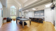 Located at 5707 Luxemburg St in Rockville, MD, this single-floor community lounge is a well-appointed gathering space designed for both relaxation and entertainment. The room immediately impresses with its soaring vaulted and coffered ceilings, which lend an airy, open atmosphere throughout. Warm hardwood flooring flows seamlessly across the entire space, complementing the bright, neutral white walls.

Natural light pours in through tall arched windows and a dramatic floor-to-ceiling window alcove, where two full blue sofas face each other around a round ottoman — creating an inviting conversation nook with serene tree-lined views. Skylights overhead further enhance the bright, welcoming ambiance.

A centerpiece pool table with a sleek modern frame anchors the main area, offering a focal point for recreation. Adjacent to it, a dark-finished bar counter with a white quartz top is flanked by wood-topped bar stools and accented by a striking blue subway tile backsplash and pendant lighting — ideal for casual gatherings. A wall-mounted flat-screen TV adds to the entertainment options nearby.

The space also features colorful artwork displayed on the walls, adding personality and warmth. French doors at the rear provide additional access and bring in even more natural light, rounding out this thoughtfully designed communal space.