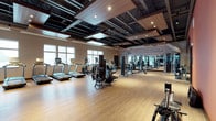 This fitness center offers a spacious, single-floor layout designed to accommodate a full range of exercise activities. The open floor plan is anchored by light hardwood-style flooring that lends the space a clean, energetic feel. Soaring ceilings with exposed structural elements and warm wood paneling are complemented by recessed lighting, creating a bright and motivating environment. Large windows along one wall invite generous natural light, enhancing the airy atmosphere throughout.

The cardio zone features a row of treadmills and elliptical machines, each equipped with individual screens for entertainment during workouts. Strength training is well represented with a variety of cable machines, resistance equipment, and free weights arranged for easy access and safe movement between stations. A mirrored wall on one side expands the sense of space while allowing members to monitor their form. A motivational typography mural stretches across a prominent wall, featuring bold phrases such as "Good Things Come to Those Who Sweat," adding an inspiring visual element to the workout environment. The thoughtful layout ensures ample floor space between equipment, supporting a comfortable and functional fitness experience.