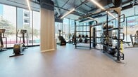 Located at 3545 Wilshire Blvd in the heart of Los Angeles, this contemporary fitness center offers a thoughtfully designed, single-floor layout spanning a generous footprint across multiple workout zones. Floor-to-ceiling windows line the perimeter, flooding the space with abundant natural light while framing sweeping views of the surrounding Koreatown skyline and lush palm trees — a distinctly Los Angeles backdrop for any workout.

The facility is organized into several distinct areas catering to a wide range of fitness activities. A dedicated cardio zone features a row of treadmills and upright bikes positioned directly facing the expansive windows, maximizing the outdoor views during exercise. A separate cycling studio houses a lineup of spin bikes on light hardwood flooring, with large glass walls maintaining an open, airy feel.

The strength training area is well-appointed with a comprehensive selection of resistance machines, free weights, weight racks, and a functional training rig complete with battle ropes and medicine balls. An exposed ceiling with industrial-style linear lighting adds a modern, urban aesthetic, complemented by warm wood-panel accent walls throughout.

Rubber and carpet flooring are used purposefully across zones to suit each activity. Mirrored walls in the strength area enhance the sense of space. The overall ambiance is sleek, bright, and motivating — a premium fitness environment in the heart of one of Los Angeles' most vibrant corridors.