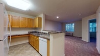 This single-floor residential unit offers a functional and well-organized layout designed for comfortable everyday living. The kitchen is equipped with warm oak cabinetry, dark laminate countertops, a white electric range with overhead ventilation, a dishwasher, and a double sink. A peninsula counter separates the kitchen from the adjoining living area, creating an open, connected feel while maintaining distinct zones for cooking and relaxing. The living area features plush wall-to-wall carpeting and is finished with neutral, warm-toned walls that lend a calm and welcoming ambiance. A window unit provides climate control, and recessed lighting illuminates the space effectively. The bedroom is generously proportioned, also finished with soft carpeting, and features a wide window fitted with blinds that frames views of mature trees outside, allowing natural light to filter in. The bathroom includes a single-sink vanity with oak cabinetry, a large mirror, tile flooring, and a tiled shower-tub combination. Throughout the unit, tile flooring in the kitchen and bathroom contrasts with the carpeted living and sleeping areas. The overall design is clean, practical, and move-in ready.